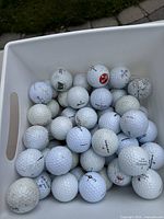 White plastic container filled with about 50 used white golf balls showing various logos including Coca-Cola, some visible dirt and scuff marks on the surface.