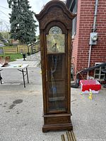 Full front view of a tall antique style grandfather clock made of brown wood with decorative carved arch at top, glass front door showing pendulum and chains.