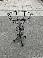 Front-left angled view of black wrought iron plant stand with twisted design on asphalt ground.