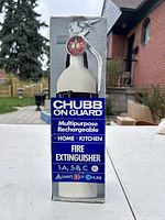 Front view of boxed white Chubb On Guard multipurpose rechargeable fire extinguisher with red pressure gauge and usage classification A 1, B 5, C.