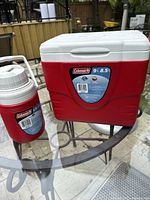 Coleman 9 Quart red cooler with white lid and accompanying red beverage jug with white top shown together on glass outdoor table.