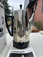 Photo of the Cuisinart cordless coffee percolator stainless steel pot with black handle and base set outdoors next to its box and instruction manual.