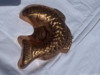 Fish shaped metal baking mold showing detailed scales, fins, and open mouth, photographed on white fabric background.