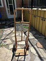 Front view of the metal utility dolly showing orange frame, two wheels, and platform, positioned outdoors on stone ground, with visible rust and wear.