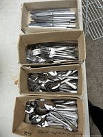 Four cardboard boxes each containing one type of flatware: knives, dinner forks, teaspoons, and soup spoons. All pieces have matching decorative handles.