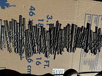 Wide angle photo showing about 60 mixed sizes of metal twist drill bits laid out in a row on cardboard. Bits show wear and surface rust.