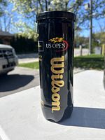 Black Wilson US Open tennis ball can with gold lettering showing the Wilson logo and US Open branding in outdoor natural daylight.