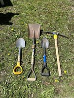 Four pre-owned gardening tools laid on grass: large Ridgid square shovel, small yellow-handled shovel, small black-handled shovel, and a pickaxe with wooden handle and yellow grip.
