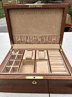 Open jewelry box showing various compartments lined in beige fabric with several ring, earring, and bracelet holders along with hooks and elastic pouch on lid.