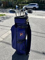 Spectra golf irons set in a blue Fila branded golf bag, showing the golf clubs with their steel shafts and club heads, outdoors on pavement near a street.