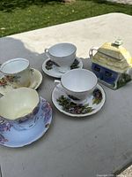 Three porcelain teacups with matching saucers and a decorative house-shaped teapot displayed on outdoor table.