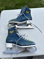 Pair of side-profile view of vintage figure skates to show denim stylized uppers, yellow patches, stacked white heels and soles, and blades.