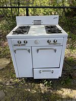 Front view of vintage white Findlay stove showing two gas burners on top, four control knobs, one cabinet door on left, oven door in center, and drawer below oven.