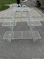 Full side view of white metal plant stand showing three rectangular wire grid shelves and side scrollwork design on paved ground outside