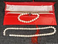 Pearl necklace and matching bracelet displayed inside a red box on a textured gray surface. Necklace and bracelet have a string of white pearls.