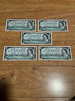 Five Canadian one dollar banknotes laid flat on wood flooring, showing front sides with Queen Elizabeth II portrait and centennial dates 1867-1967.