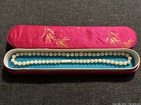 Pearl necklace displayed in open red ornamental box with gold leaf embroidery and blue fabric interior.
