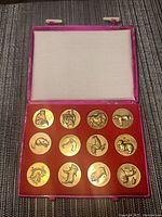 Photo showing the closed pink case with 12 gold-colored Chinese zodiac coins arranged in two rows of six each on a red velvet insert.