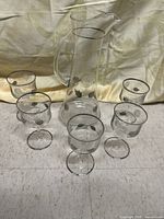 Full view of the vintage glass pitcher and all five matching glasses arranged around it, showing silver leaf decoration and silver trim on clear glass.