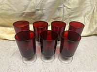 Seven ruby red vintage glasses displayed from a frontal angle showing their red colored bodies and clear bases on a light surface.