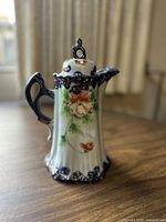 Porcelain chocolate pot decorated with hand-painted orange flowers, green foliage, royal blue base and handle, gold accent trim, displayed on wooden table.