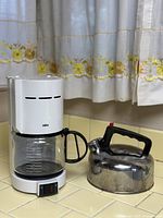 Photo of Audi Braun Aromaster Classic drip coffee maker with glass carafe and stainless steel teapot kettle on yellow countertop in kitchen setting with floral curtains.