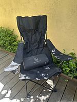 Full view of black Mercedes-Benz folding chair set up outdoors on wooden surface next to greenery. Chair fabric shows some light shading but no visible damage.