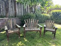 Full set of three vintage Adirondack style wooden chairs positioned outdoors on grass with greenery and wooden fence in background.
