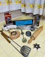 Assorted vintage kitchen accessories including garlic press, wooden rolling pin, cookie cutters, potato masher, egg slicer, meat tenderizer, and spatula laid out on a tiled kitchen counter under yellow floral curtains.