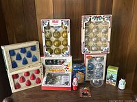 Vintage Christmas decorations including boxed glass ornaments, string lights, and festive figurines arranged on a wood-paneled background.