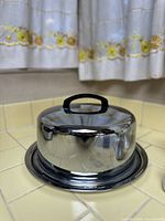 Chrome metal cake carrier with dome lid and black handle, placed on beige tiled kitchen counter in front of curtains with yellow floral embroidery.