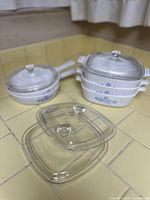 Five vintage CorningWare casserole dishes stacked with matching glass lids placed nearby on the counter.