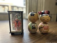 Lot showing two Sosaku Kokeshi wooden dolls (male and female) side by side with a glass shadowbox containing a Geisha figurine