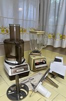 Photo showing three vintage kitchen appliances: Sunbeam food processor, cream-colored blender, and white meat slicer with attachments arranged on a kitchen counter below a window with floral curtains.