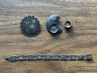 Top-down photo showing all four jewelry pieces on a wooden surface: llama pendant brooch, floral brooch, ring, and bracelet with intricate gold-tone designs.