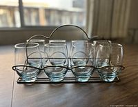 Front view of eight clear glass tumblers arranged in a gold-tone metal caddy with curved handle on a wooden surface.