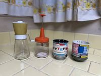 Four vintage kitchen tools arranged side by side on a tiled countertop: a manual grinder with brown lid, an orange and clear plastic chopper, and two retro printed flour sifters or measuring cups.