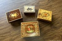 Top-down view of four vintage wooden jewelry music boxes showing various decorative tops including floral and figurative artwork.