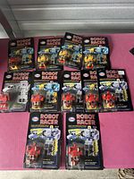11 vintage Esso Robot Racer transformers on original 1980s blister cards in a layout on a pink surface. Several yellow, red, blue, and silver robot racers visible with packaging and labels.