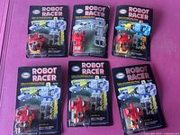 Six 1980s Esso Robot Racer toys sealed on original purple-backed cards with yellow and white text, showing front view of robotic transforming vehicles in red, blue, and yellow plastic.