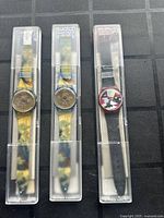 Three vintage Swatch watches displayed in original transparent plastic cases, showing the fronts and straps of each watch.