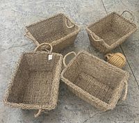 Four rectangular woven baskets with natural fiber handles, two handles need reattachment, and a rope knot door stop placed on a concrete floor.