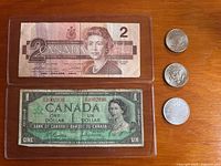 Two Canadian paper banknotes, $2 and $1, shown in protective sleeves alongside three coins on wooden surface.