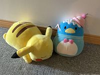 Two large squishmallow plush toys, a yellow Pikachu lying on its side and a blue penguin with cupcake hat, placed on floor near wall