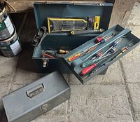 Photo showing two metal tool boxes; one large green box with a dent and one smaller gray box. The green box's removable tray is visible, filled with various hand tools including screwdrivers, scissors, a hand plane, and chisels.