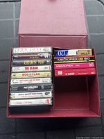 Open red storage case showing 9 vintage rock cassette tapes on left and 4 blank recordable cassette tapes stacked on right
