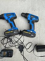 Two blue Mastercraft cordless lithium-ion screwdrivers with attached batteries and two chargers with cords laid on concrete surface.