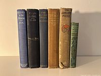 Front-facing view of 6 antique/vintage hardcover books standing upright showing spines with titles and authors, colors ranging from blue, black, brown, tan, cream, and green.