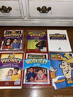 Six educational books and charts on the floor showing covers with children and cartoon characters. Includes phonics, visuals, math, and worktext materials for Kindergarten K5 level.