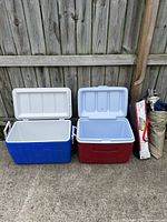 Two large hard plastic coolers, one blue and one red, with open white lids, alongside a green Coleman tent packed in its carry bag.